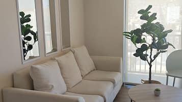 Uma sala de estar moderna com um sofá bege, uma mesa redonda de madeira e uma janela ampla com vista para edifícios.