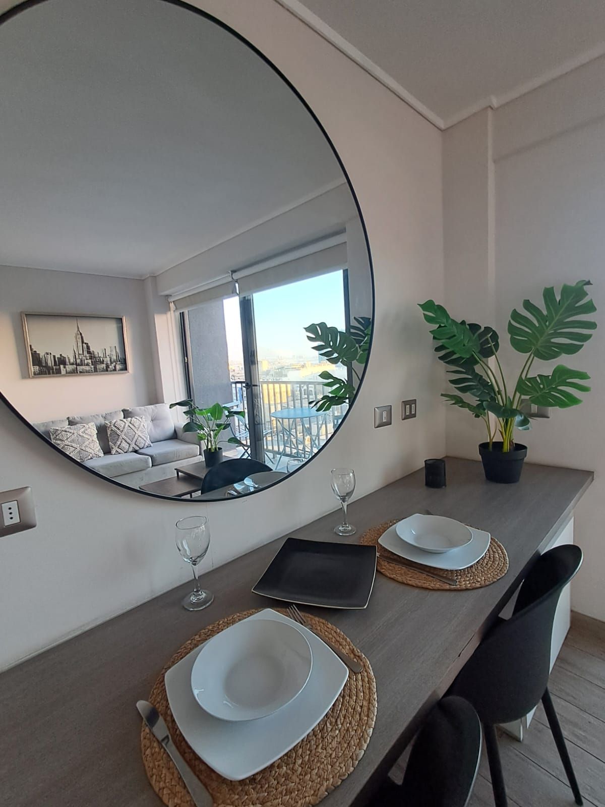 Un comedor moderno con un espejo grande que refleja un balcón y un paisaje urbano, una mesa de madera con individuales y una planta en maceta.