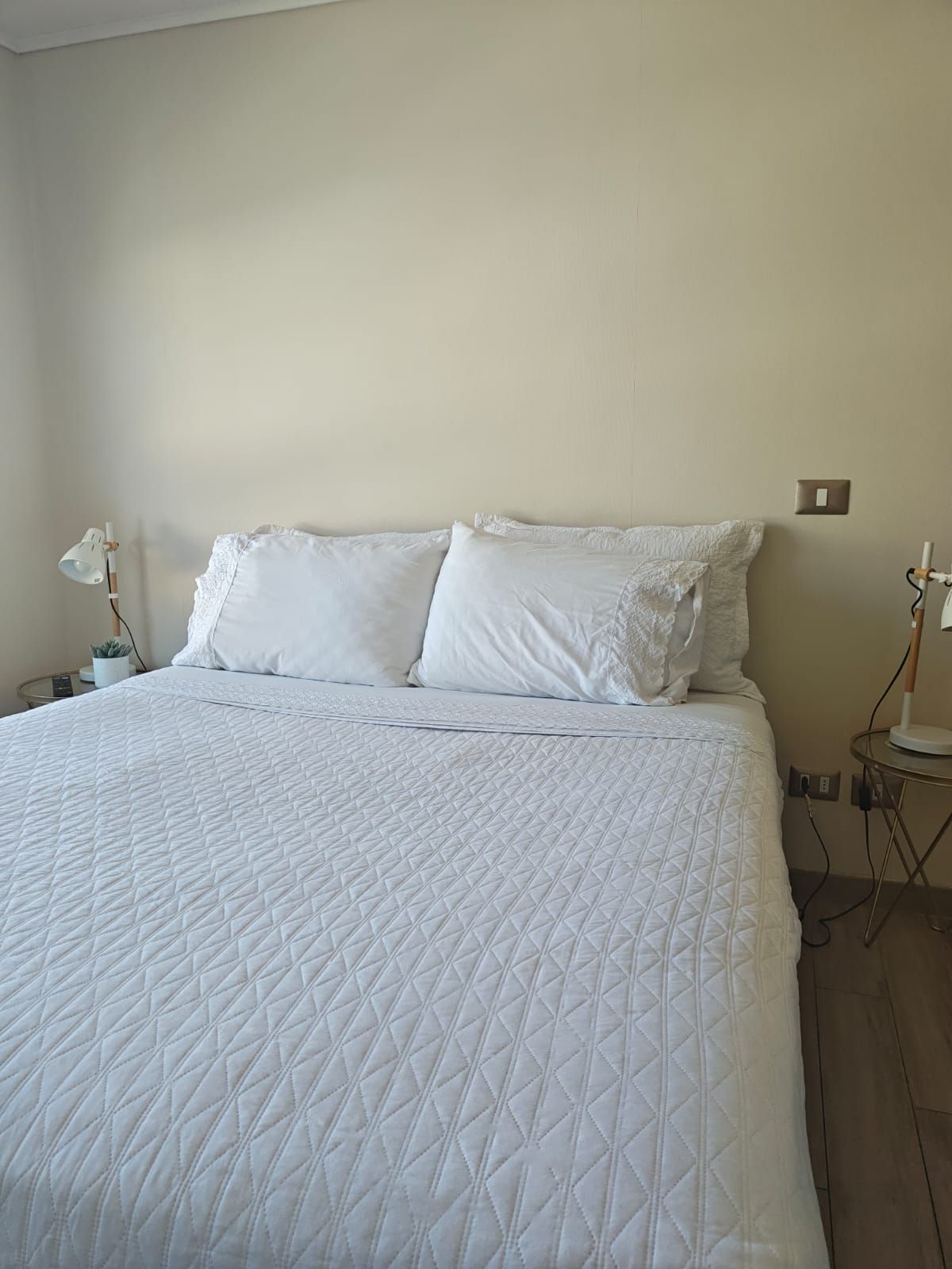 Una cama bien hecha con un edredón blanco acolchado, dos almohadas y una mesita de noche con una lámpara.