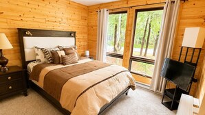 A bedroom with a wooden paneled wall, a bed with a headboard, a nightstand, a TV stand, and a window with curtains.