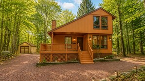 A two-story wooden cabin with a covered porch, a visible garage, and a forested backdrop.