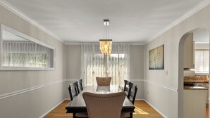 Une salle à manger avec une table en bois, six chaises, un lustre et une vue sur la cuisine, accessible par une arche.