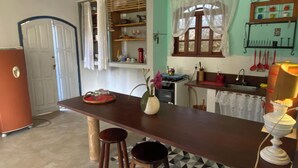 A kitchen with a wooden counter, stools, a refrigerator, a window with curtains, and a tiled floor.