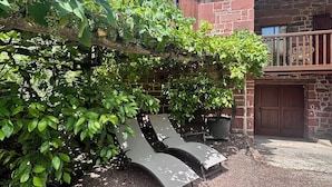 Un edificio de piedra con un balcón, una pérgola con parras y dos bancos de metal.