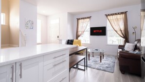 Un salon moderne avec un coin cuisine blanc, un canapé en cuir marron, une table basse en verre et un téléviseur fixé au mur qui affiche « NETFLIX ».
