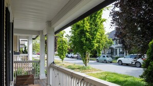 Une véranda couverte, dotée d’un banc et d’une balustrade en bois, donnant sur une rue bordée d’arbres et où sont garées des voitures.