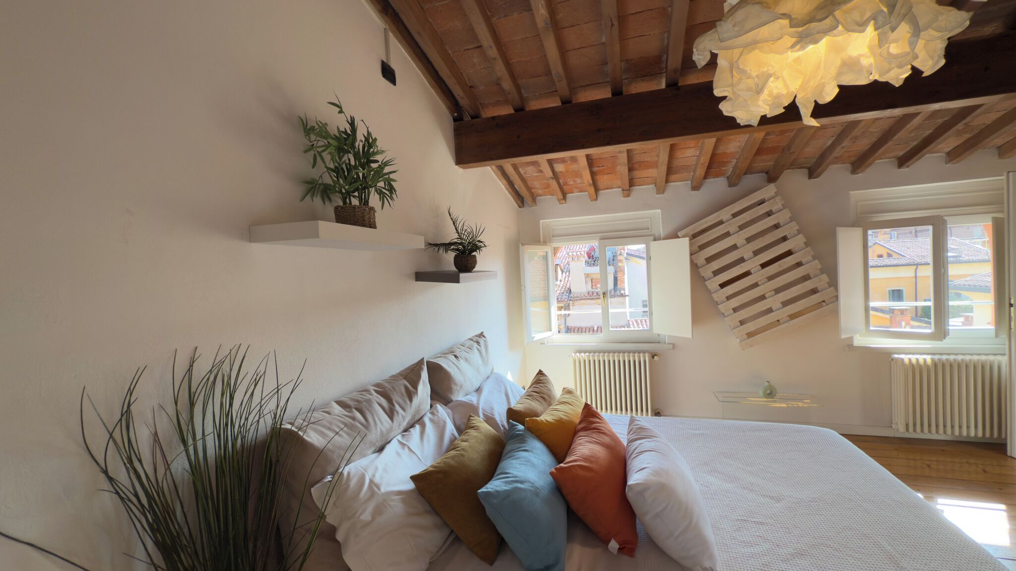 A bedroom with a bed, pillows, a hanging plant, and a view of the outside through windows.