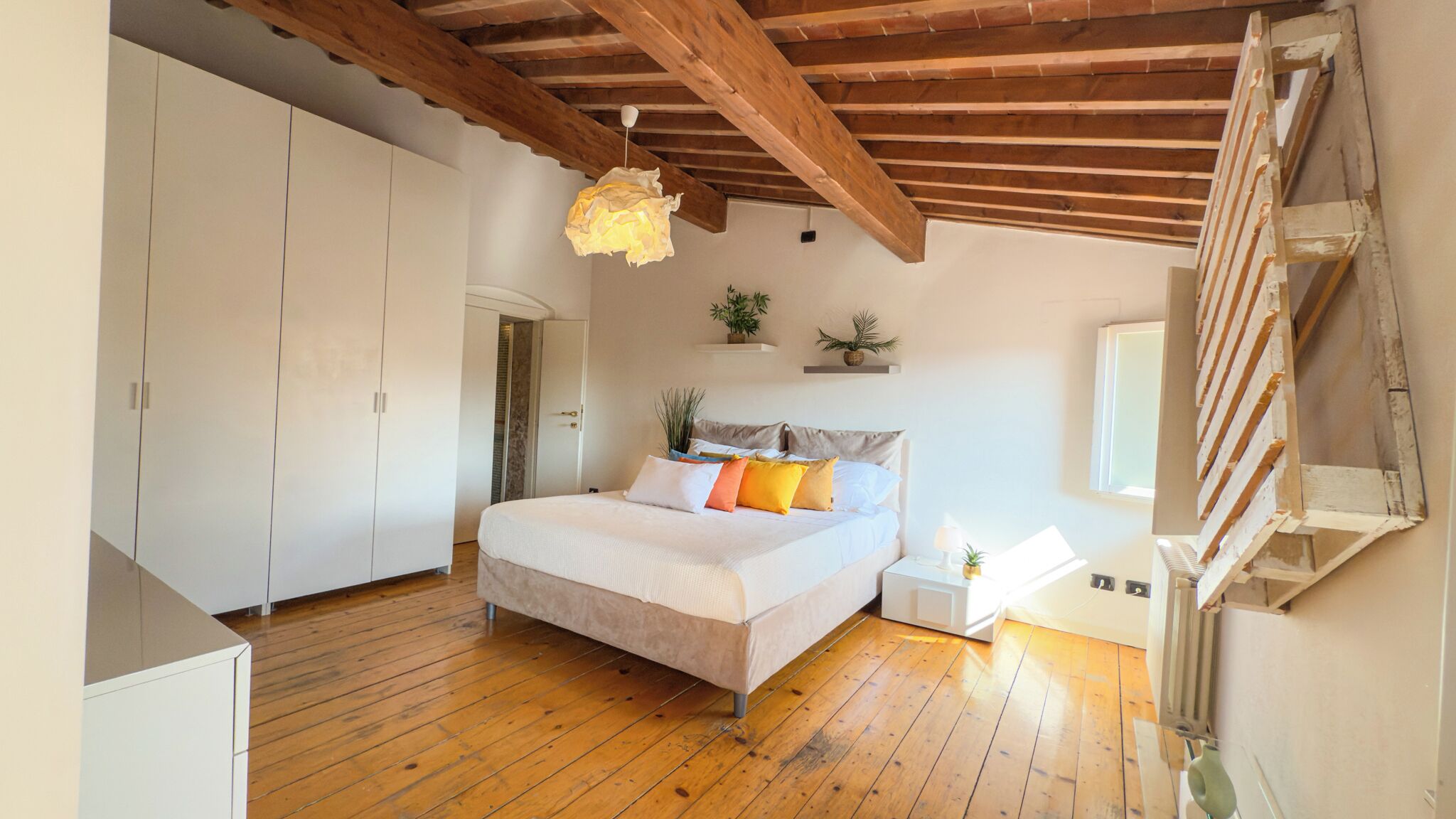 Una camera da letto con pavimento in legno, un letto con molti cuscini, una lampada a sospensione e una finestra.