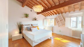 A bedroom with a wooden ceiling, a bed with multiple pillows, a bedside table, a hanging light fixture, a window with a view, and a radiator. - [Piazza Erbe] Rooftop free parking (Verona)