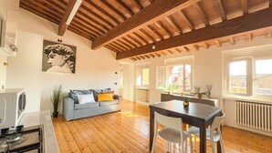 Een ruime woonkamer met houten plafond, een grijze bank met kleurrijke kussens, een eettafel met stoelen en een stadszicht door grote ramen.