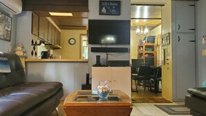A living room with a brown leather couch, a wooden coffee table with a vase, a flat-screen TV, and a kitchen area in the background.