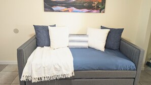 A room with a grey sofa bed, white and blue pillows, a white throw blanket, and a painting of a mountain landscape on the wall.