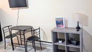 A small, neatly arranged room with a round table and two chairs, a flat-screen TV mounted on the wall, a framed welcome sign, a lamp, and a shelf with plants.