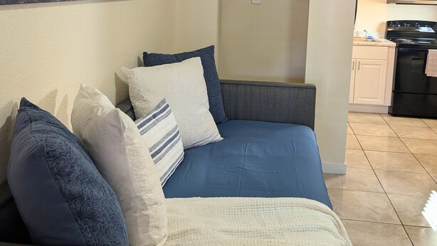A living room with a blue sofa, pillows, a painting of a landscape, and a kitchen in the background.