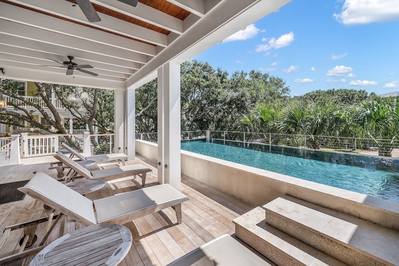 Un espacio exterior techado con piscina, sillones de descanso y vista a árboles y un edificio al fondo.