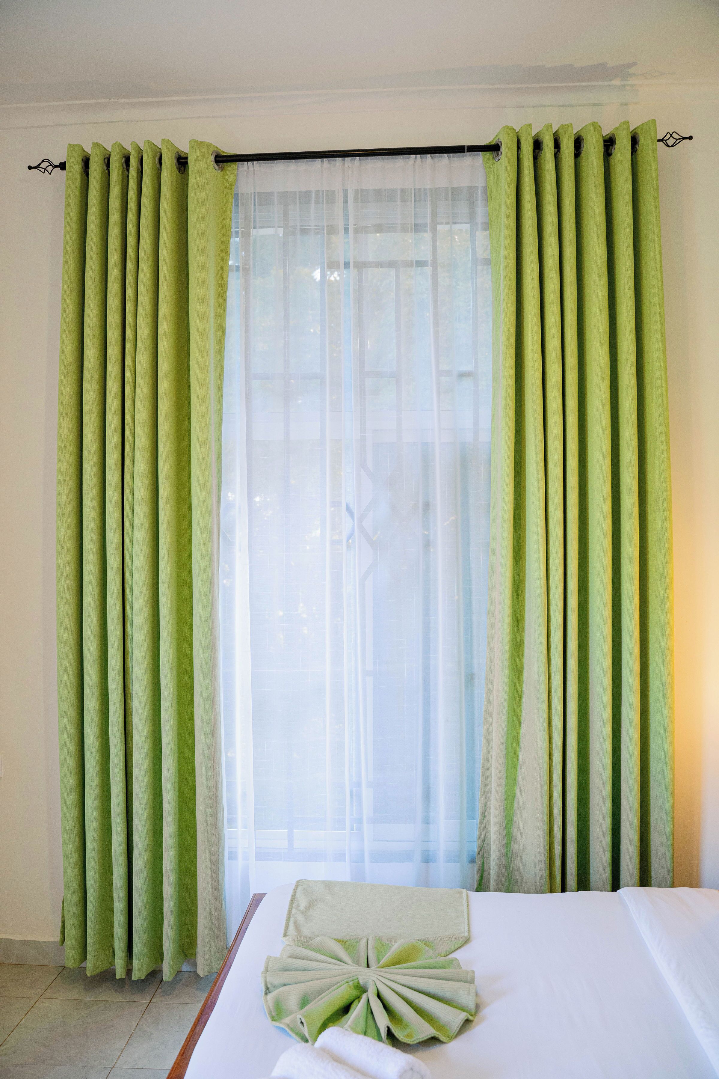 A hotel room with a bed, green curtains, and a window with sheer curtains.