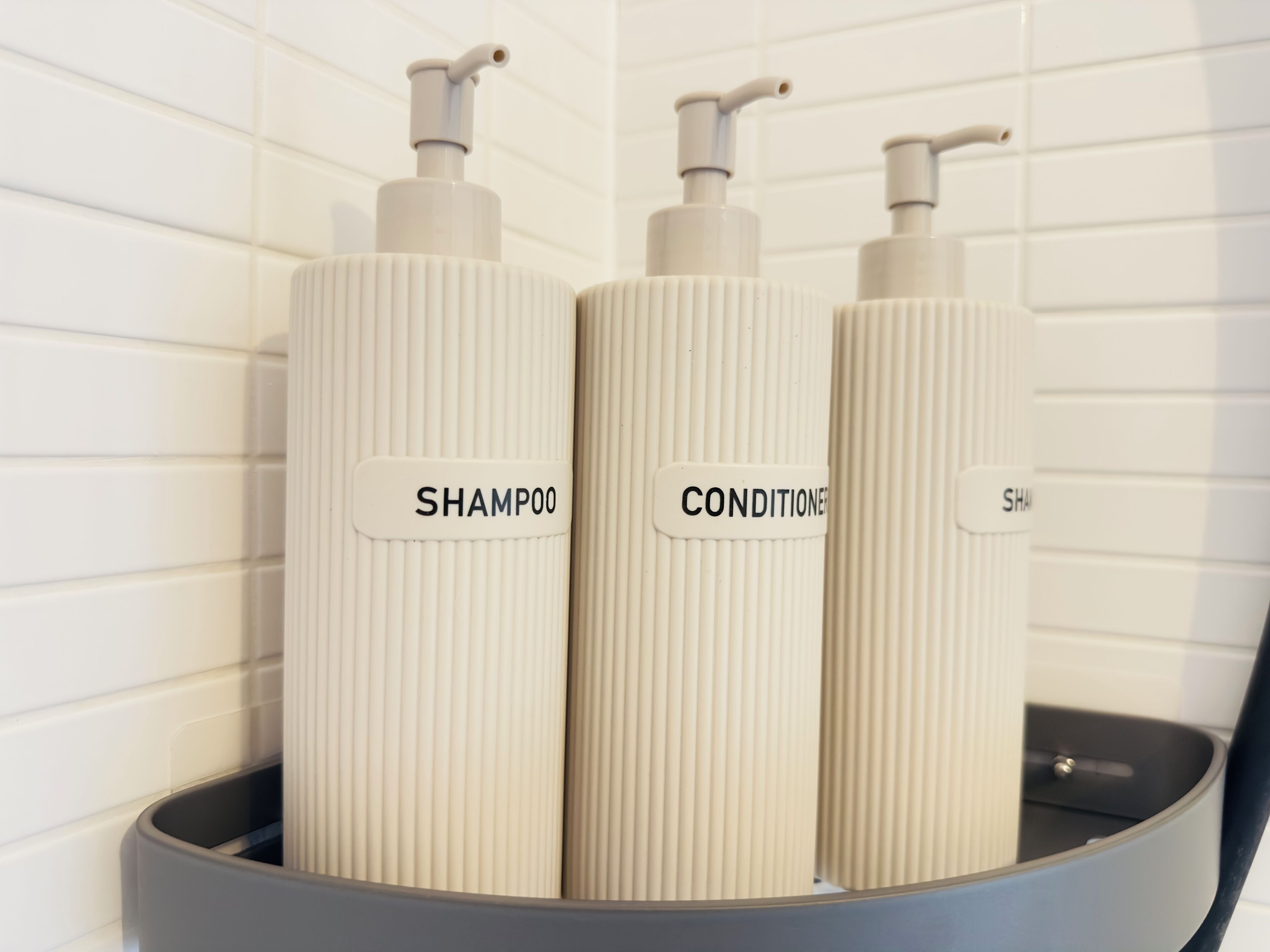 Three toiletry bottles with pumps, labeled 'SHAMPOO,' 'CONDITIONER,' and 'SHAM,' placed in a black container.