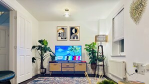 A living room with a TV, two potted plants, a floor lamp, and a remote control on a table. - Specious & Cosy 3 Bedroom House in Colchester/pet-friendly with free parking (Colchester)
