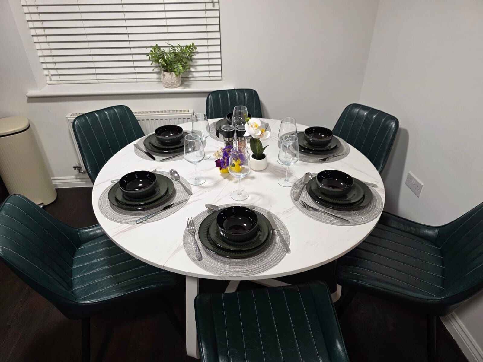 Une table de salle à manger ronde, avec de l’assiette noire, des couverts en argent et des verres à vin transparents, le tout disposé pour six personnes.