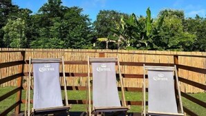 Three wooden lounge chairs with white cushions and a Corona logo, positioned on a wooden deck with a bamboo fence and lush greenery in the background. - Pousada Terras Verdes (Fernando de Noronha)
