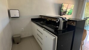 A compact kitchen with a black countertop, a white refrigerator, a small trash bin, and a wall-mounted TV. - Pousada Terras Verdes (Fernando de Noronha)