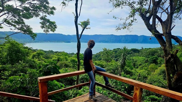 A person standing on a wooden deck with a railing, overlooking a lush green landscape and a body of water. - Cabin with beautiful view of Coatepeque Lake, Santa Ana to relax, AC, hot water+ (Santa Ana)