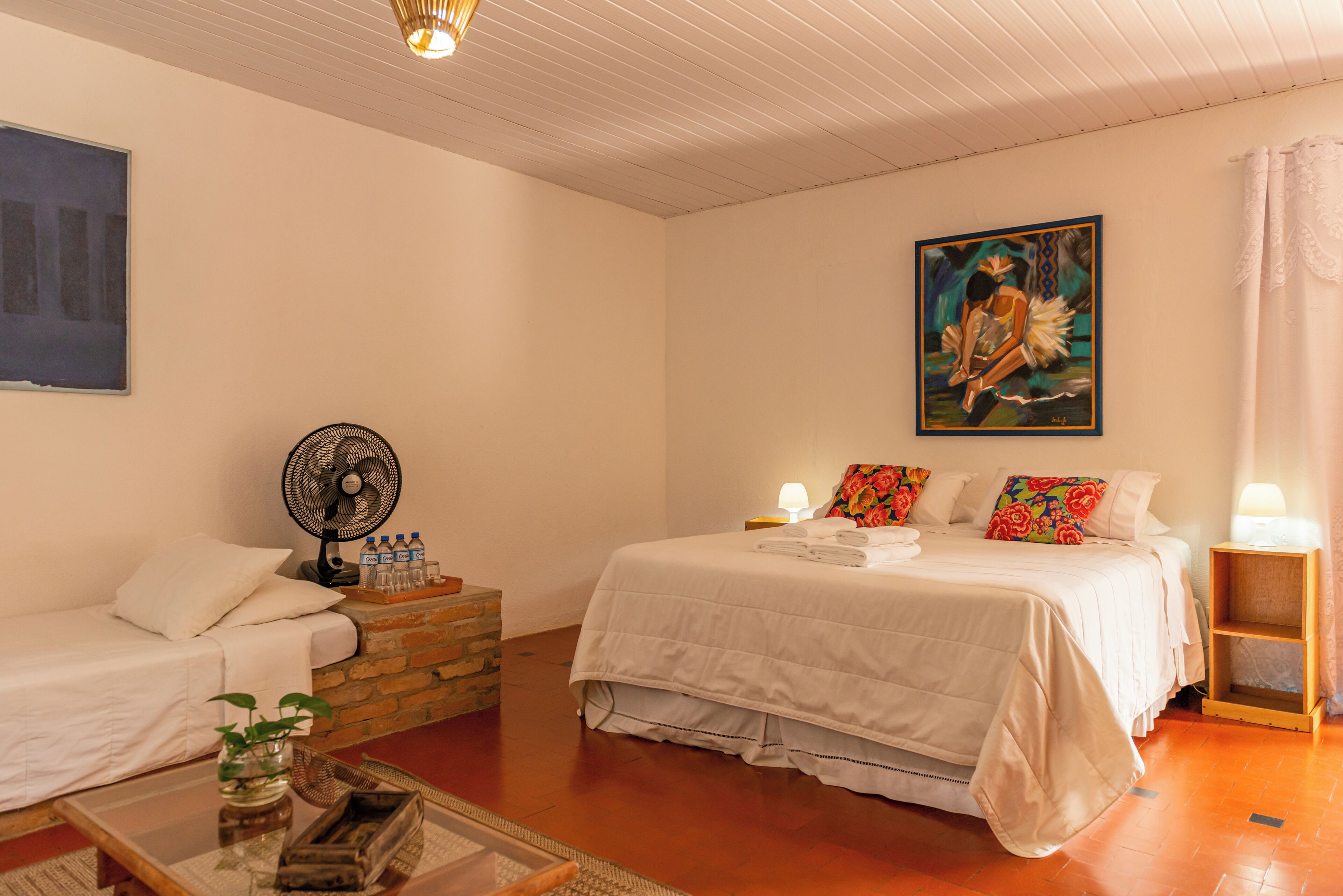 A bedroom with a bed, a sitting area with a chair and a table, a fan, and a painting on the wall.