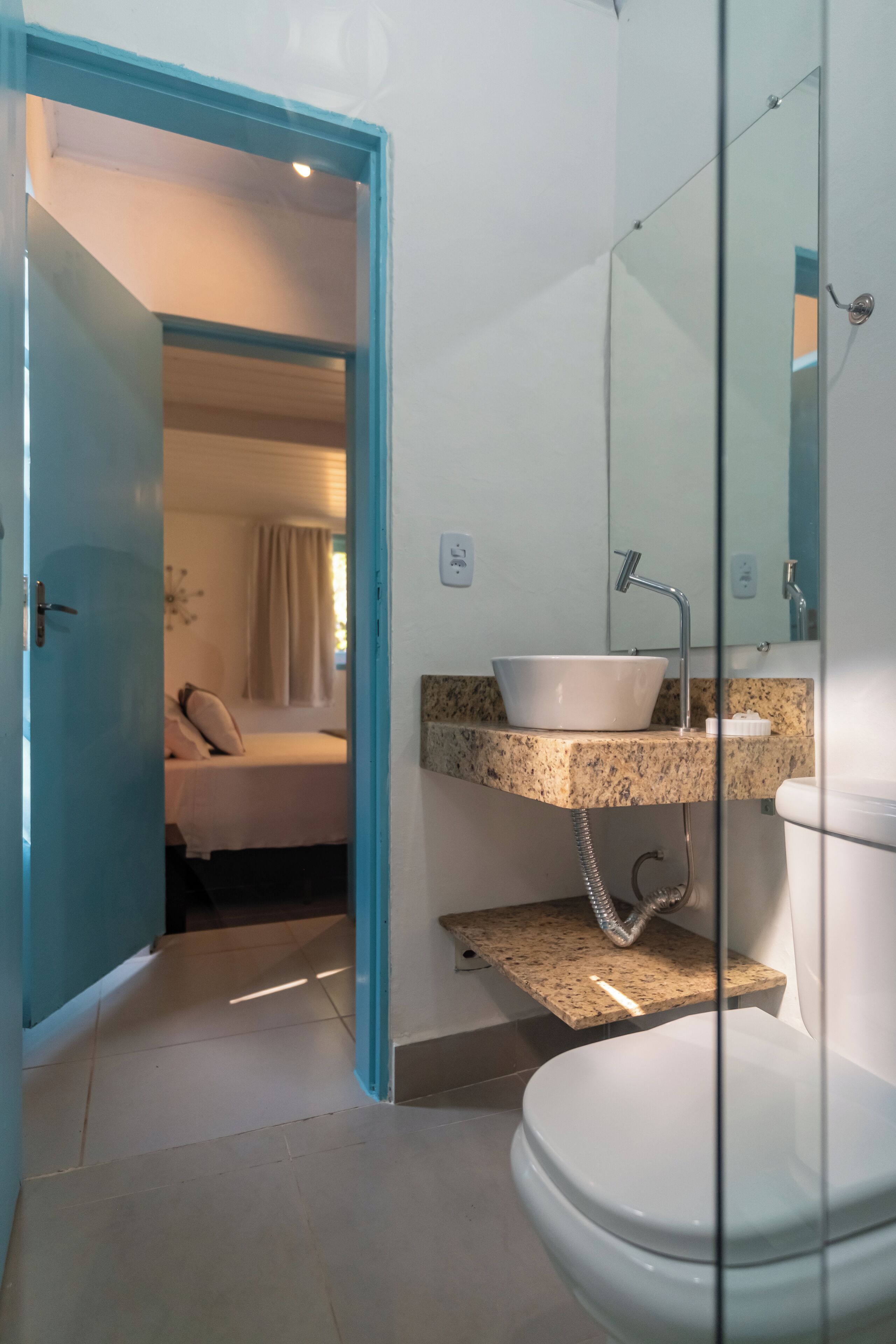 Un baño con un lavabo blanco, un espejo y un inodoro. Hay una puerta que da a un dormitorio.