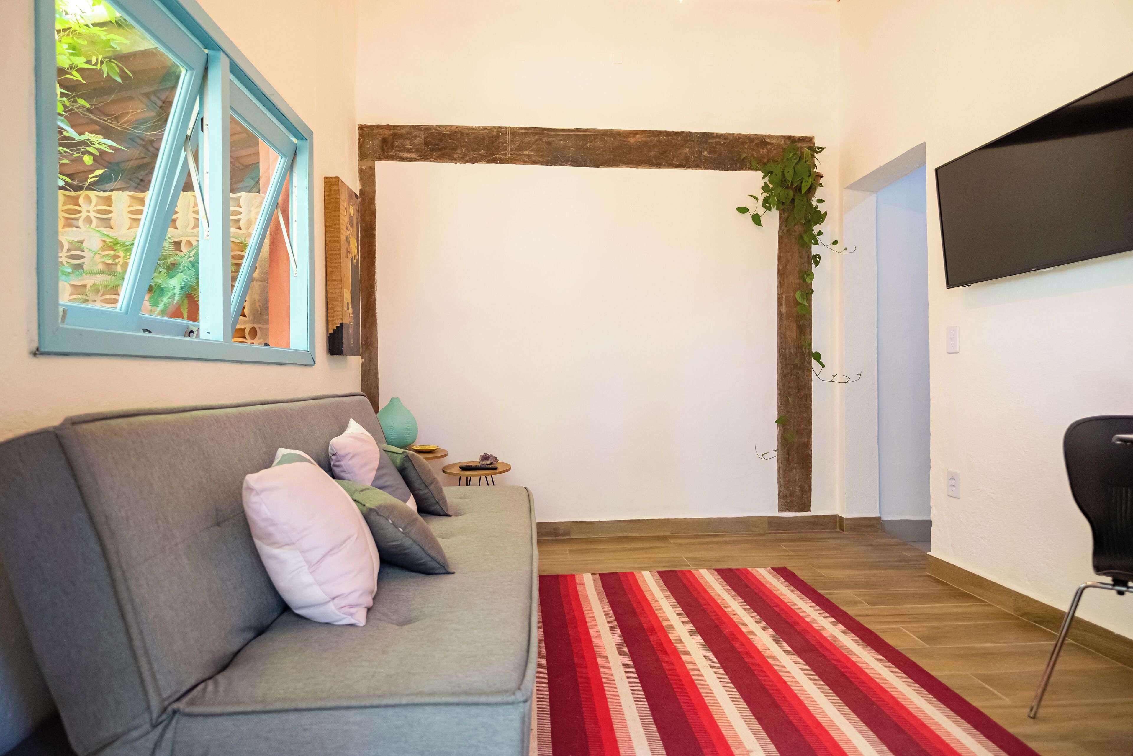 Uma sala de estar com um sofá cinza, um tapete listrado em vermelho e branco, uma TV fixada na parede e uma porta de madeira com uma planta.