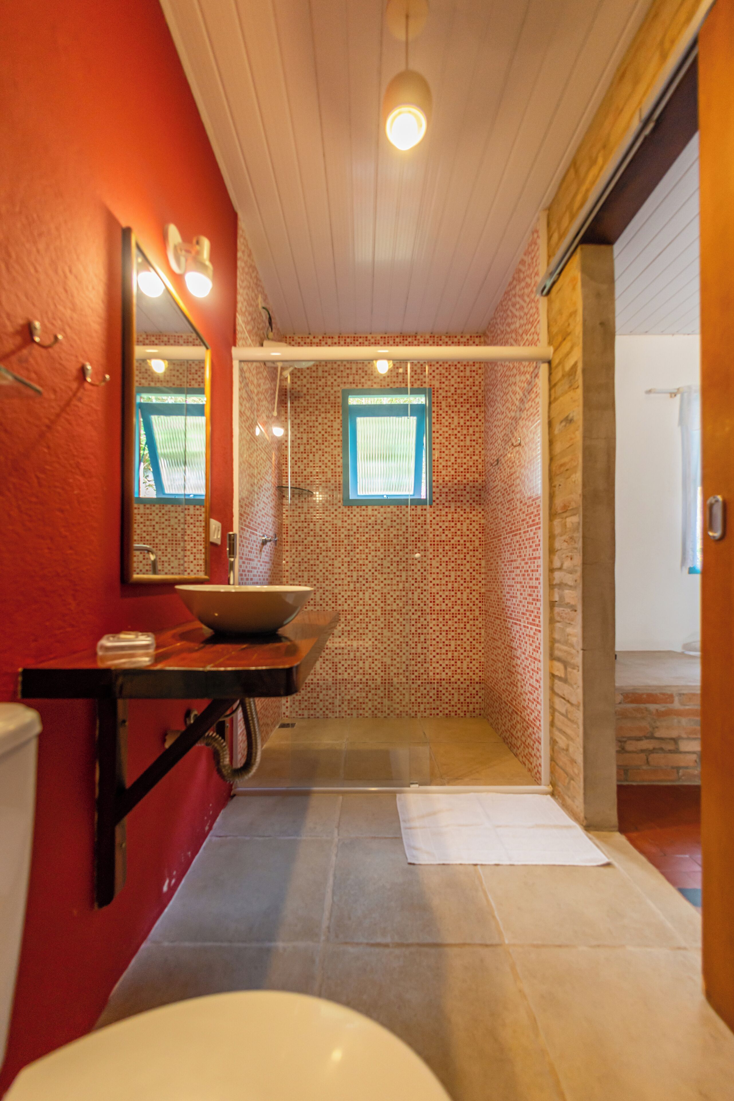 A bathroom with a red wall, a wooden vanity, a large mirror, a sink, a shower, and a window.
