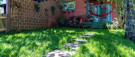 Un jardín con cerca de madera, un edificio rojo con puerta azul y una hamaca.