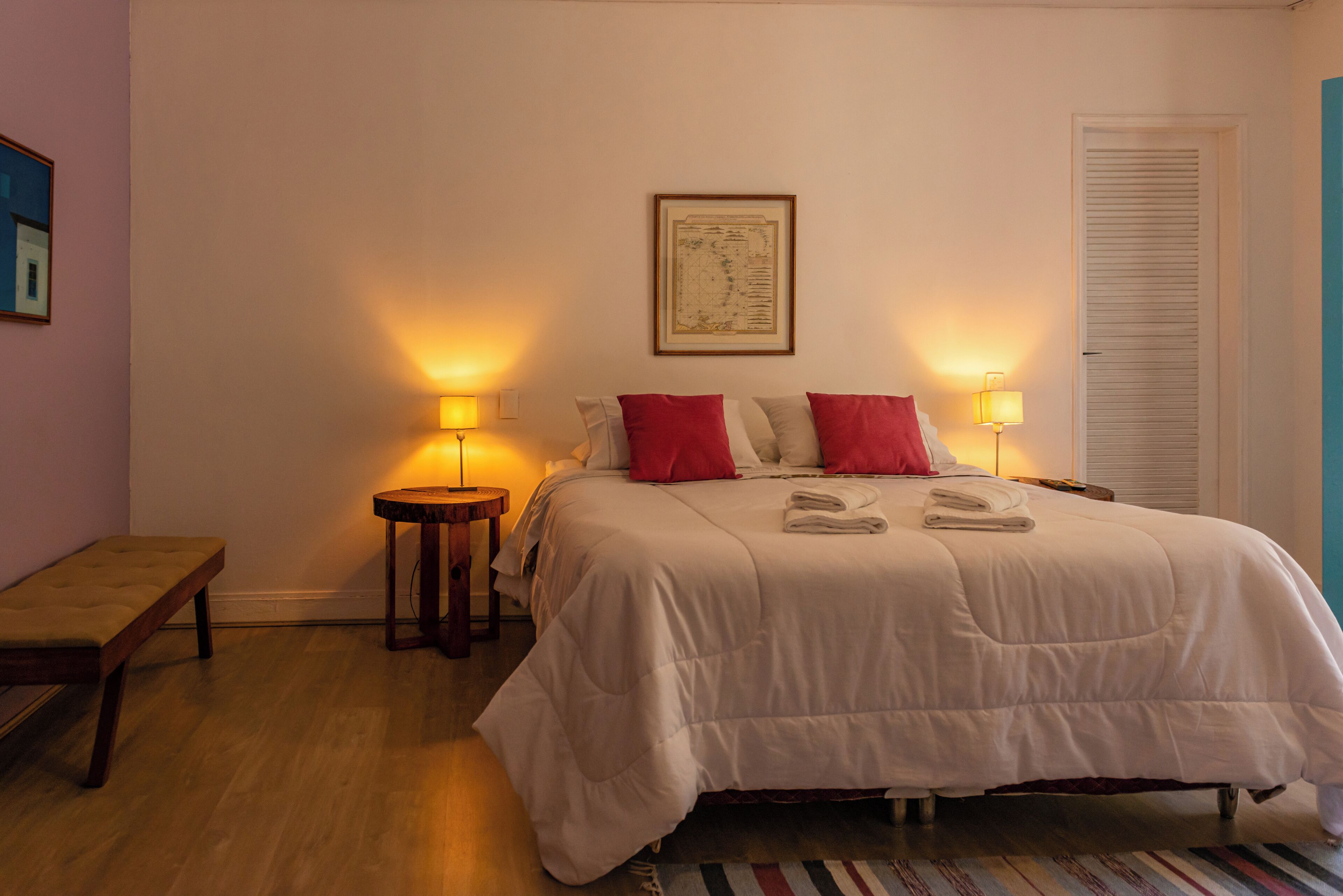 A neatly made bed with red and white pillows, two bedside lamps, a framed map on the wall, and a bench in the corner.
