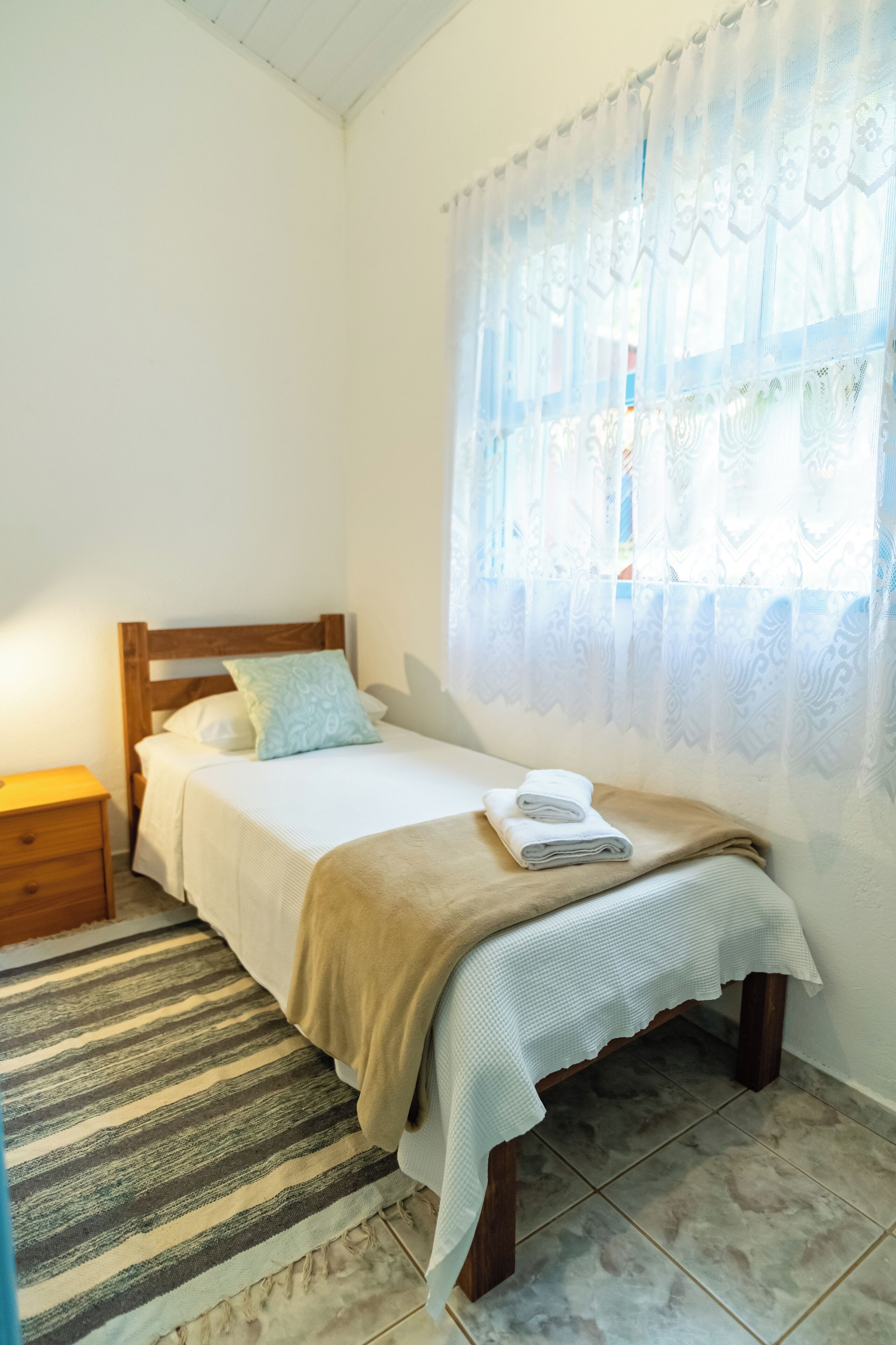 Una cama bien tendida con un tapete a rayas, una mesita de noche de madera y una ventana con cortinas de encaje.