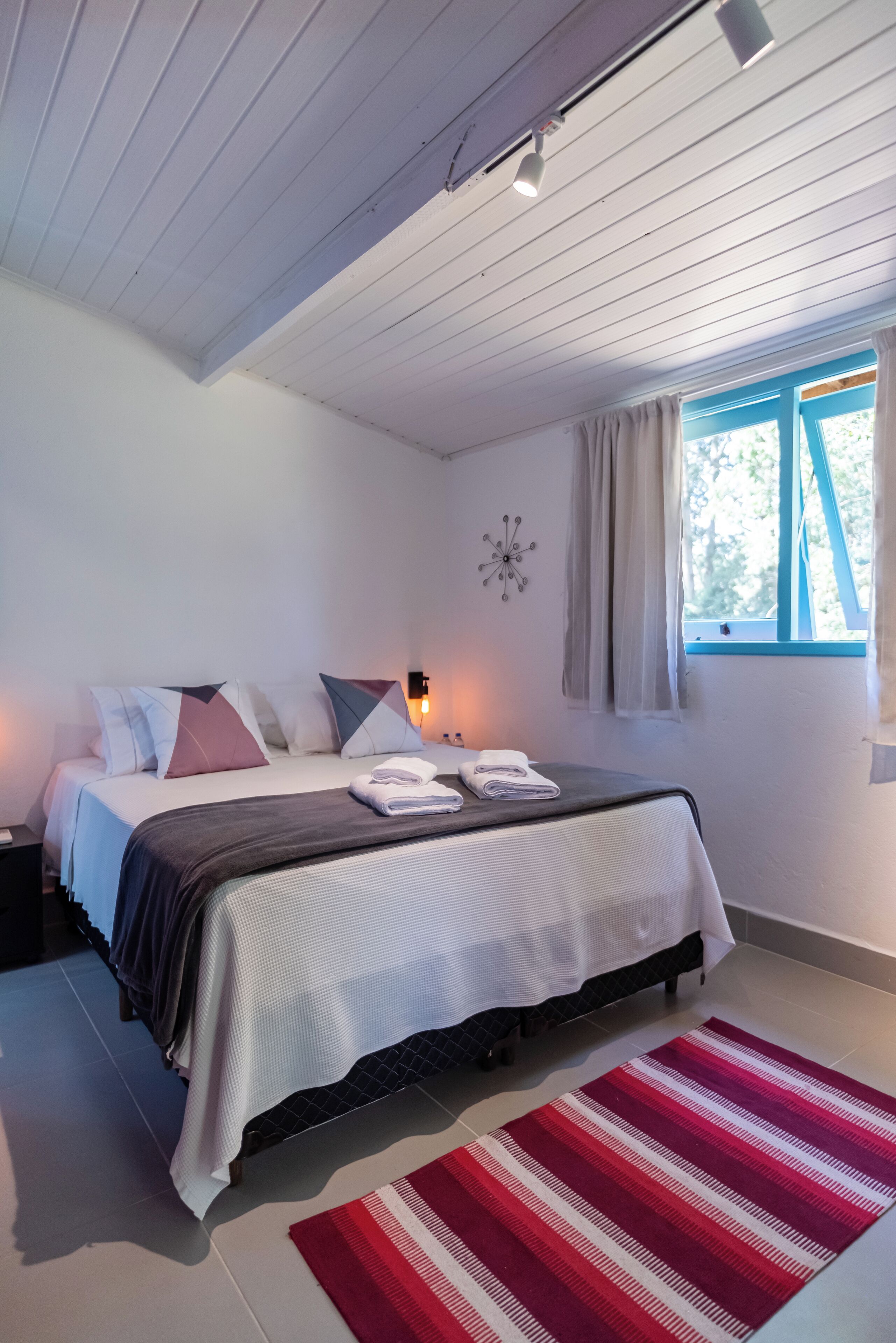 A bedroom with a bed, pillows, a nightstand, a wall clock, a window with curtains, and a red and white striped rug.