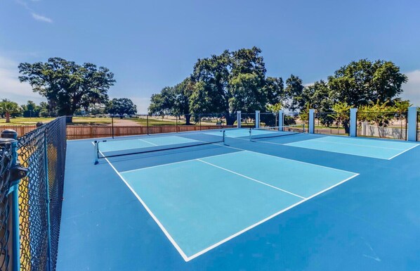 Una cancha de tenis al aire libre con red, rodeada de árboles y una valla.