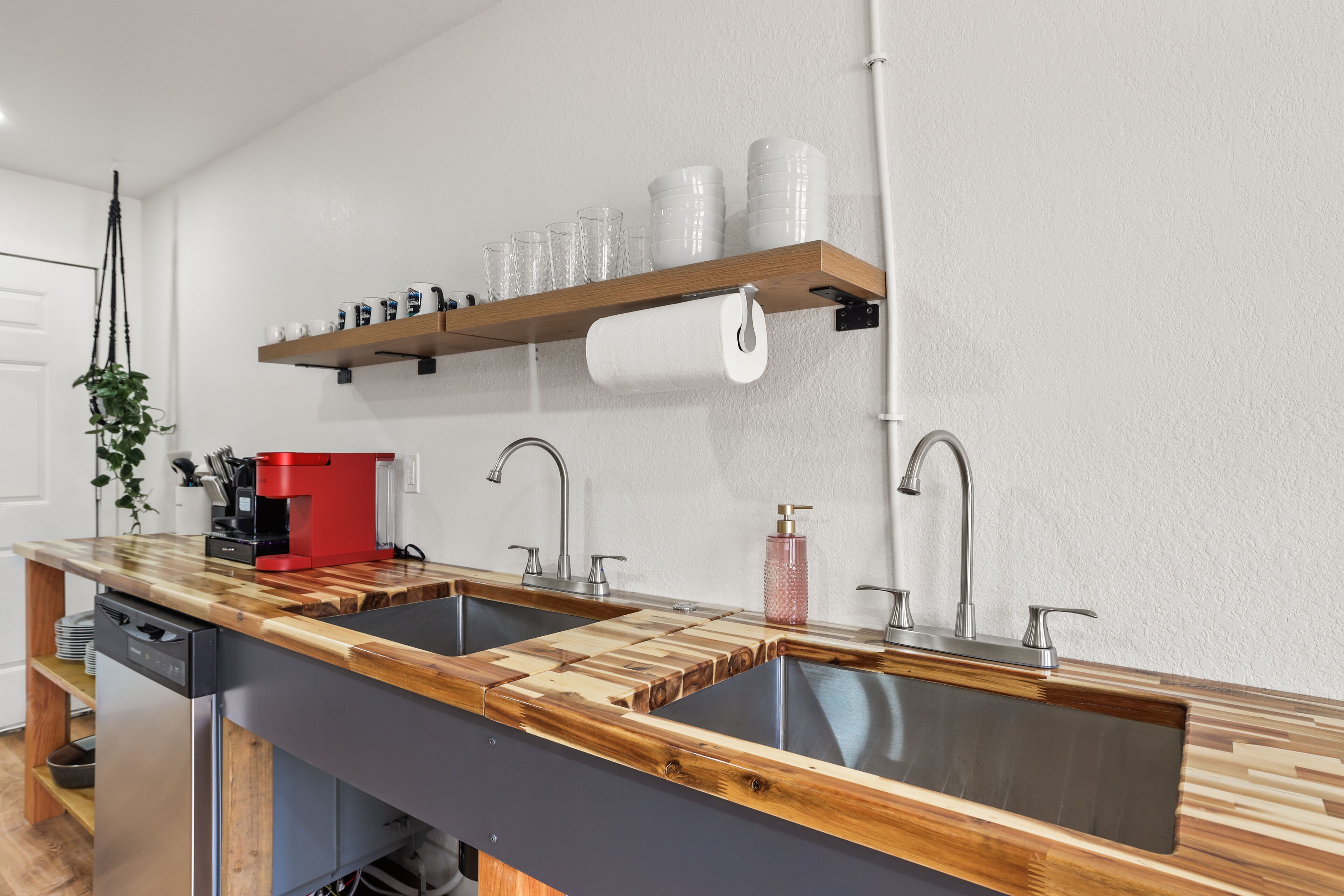Una cocina con dos fregaderos, encimeras de madera, una cafetera y un estante con tazas y un rollo de toallas de papel.
