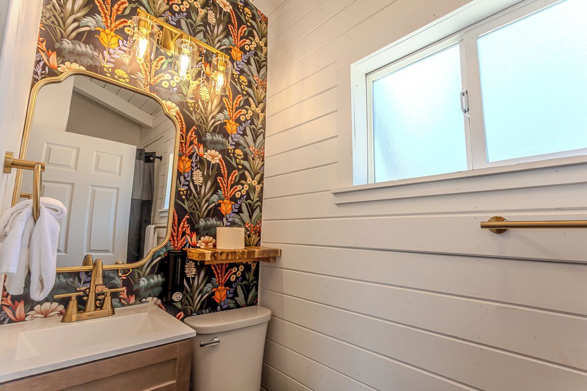 Une salle de bain avec un papier peint à motifs floraux, des toilettes blanches, un lavabo avec un robinet doré et une fenêtre située au-dessus des toilettes.
