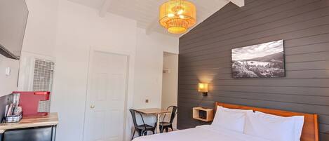 A bedroom with a bed, a desk with a chair, a small table, and a picture on the wall.