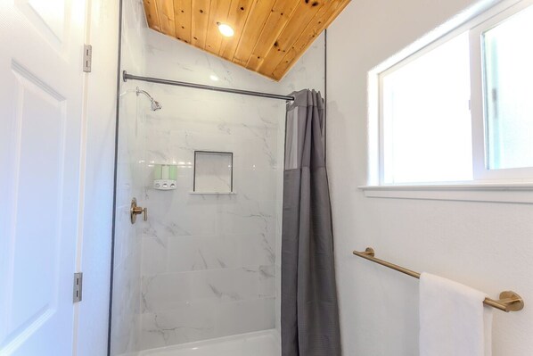 Une salle de bain moderne dotée d’une douche en verre, de carreaux de marbre, d’un plafond en bois et d’une fenêtre laissant entrer la lumière naturelle.