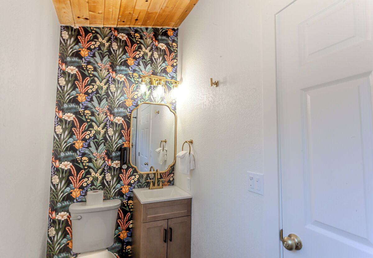 Une salle de bain avec un papier peint à motifs floraux, des toilettes blanches, un lavabo avec un robinet doré et un miroir avec un cadre doré.