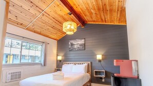 A bedroom with a wooden ceiling, a bed with white bedding, a painting on the wall, and a small desk with a red lamp. - Tahoe Mountain Inn #16 (SOUTH LAKE TAHOE)