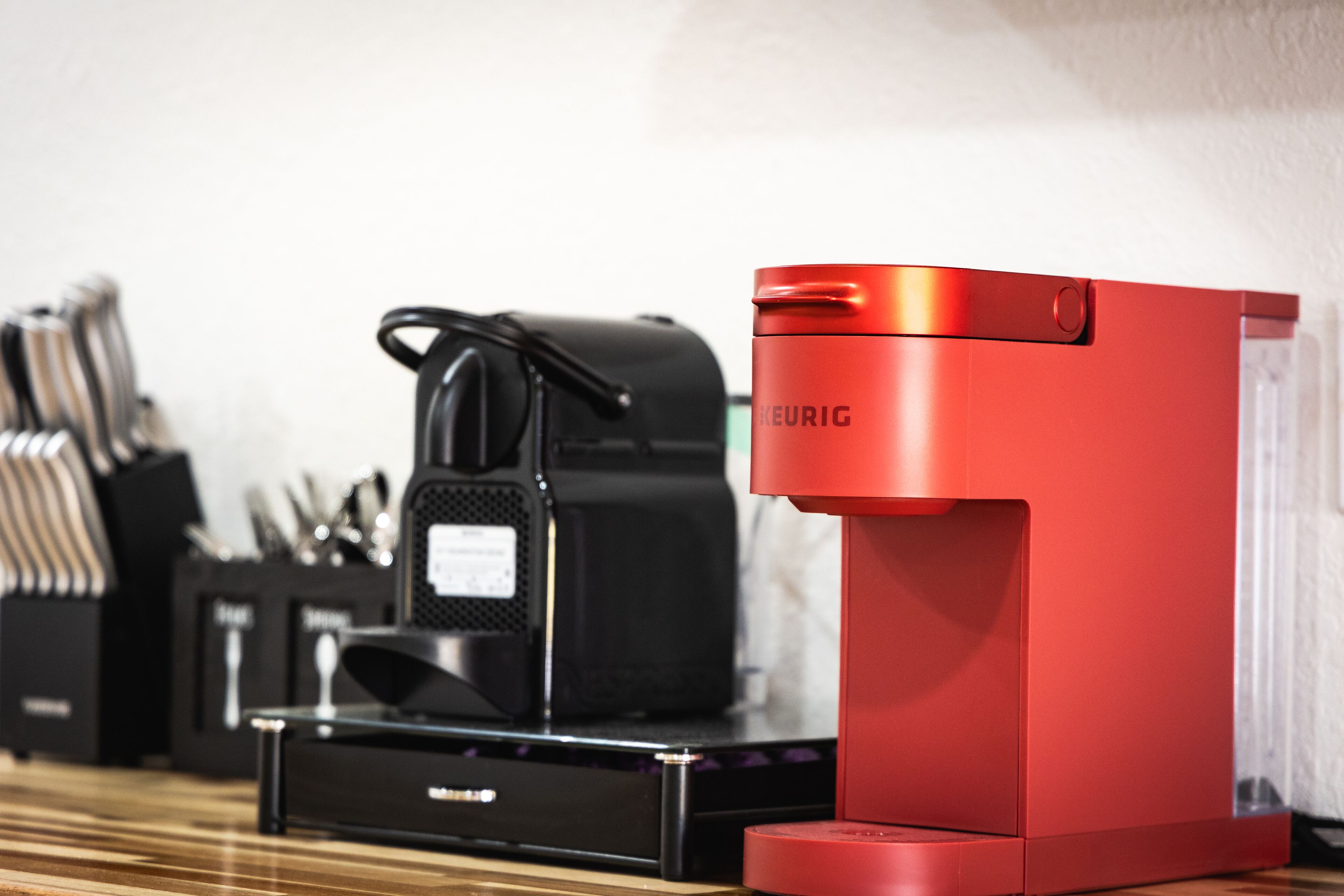 A red Keurig coffee maker on a wooden table next to a black Keurig machine.