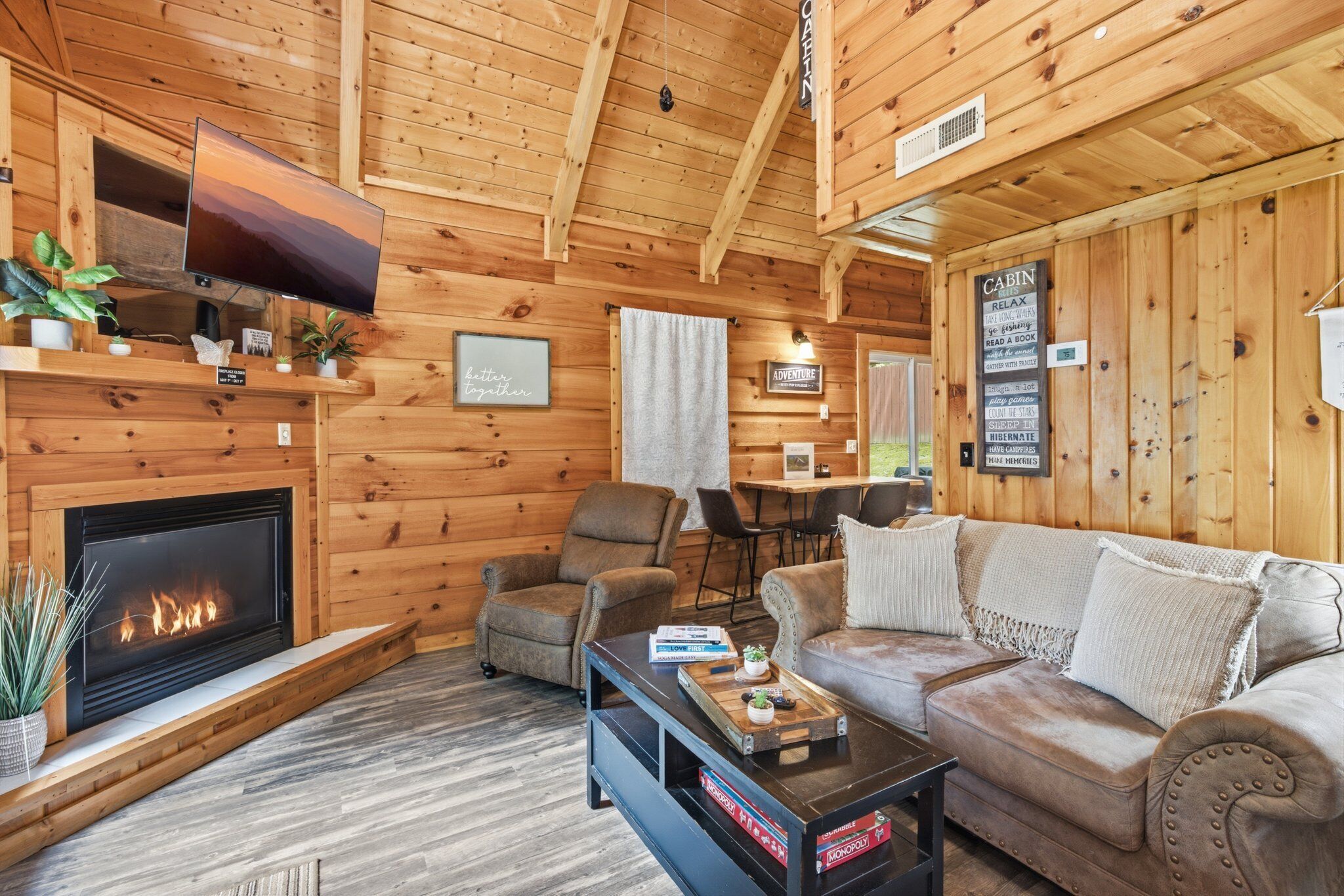 A cabin interior with a fireplace, wooden walls, a sofa, a TV, and a dining area.