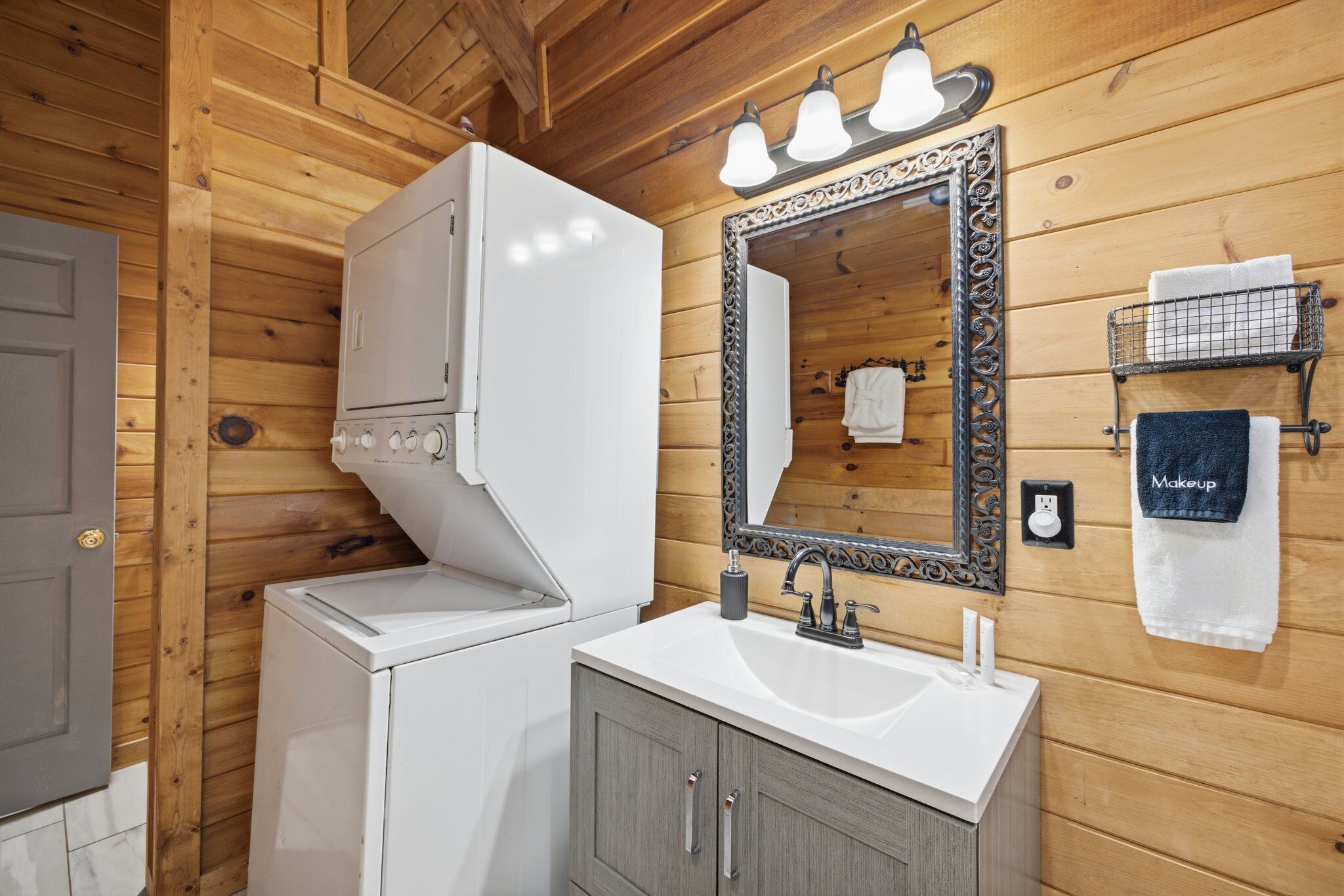 Interior de una cabaña de madera con una lavadora blanca, un lavabo con espejo y un perchero con toallas.