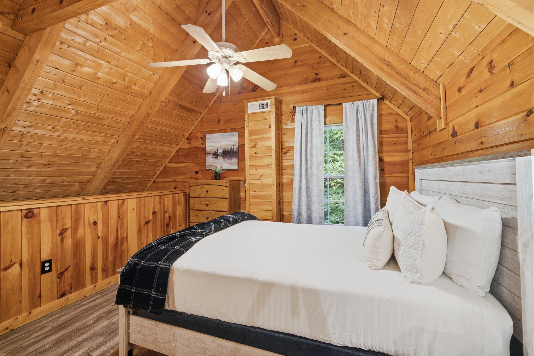 A wooden cabin bedroom with a bed, a ceiling fan, a window with curtains, and a painting on the wall.