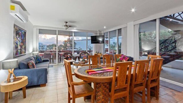 A spacious living area with a dining table, sofas, and a TV. Large windows offer a view of the outdoors. - Penthouse Unit, Great for Families (Cozumel)
