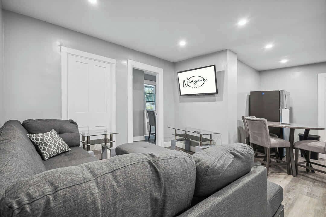 Una sala moderna con un sofá gris, una mesa de comedor con sillas y un refrigerador.