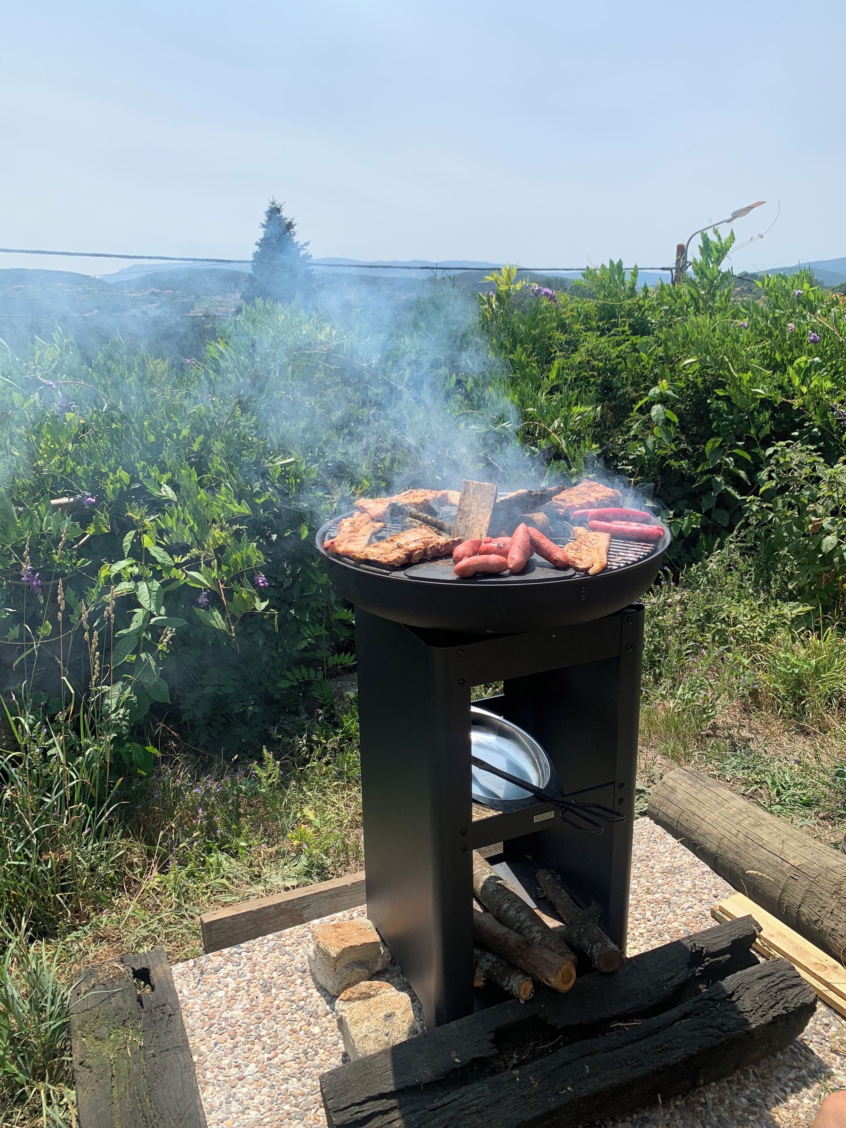 A portable grill with sausages and a steak being cooked outdoors.