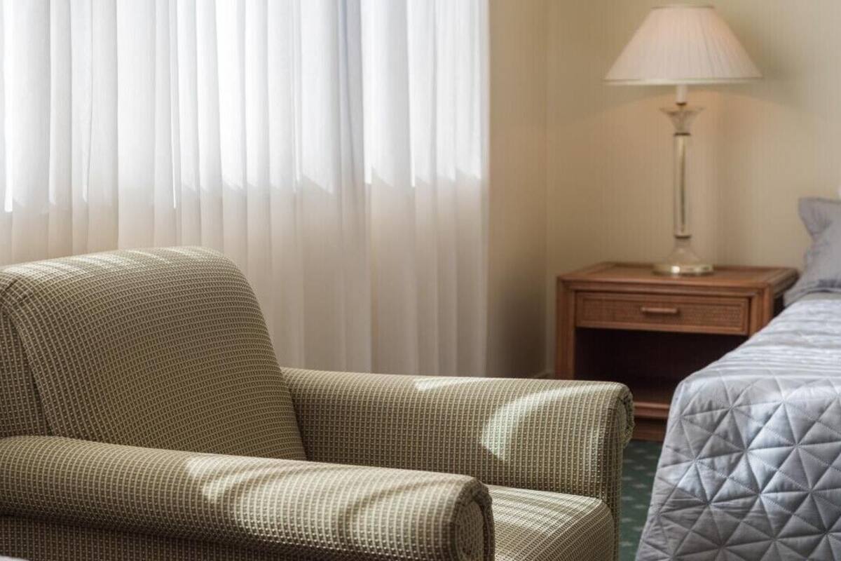 A hotel room with a single bed, a chair, a nightstand with a lamp, and sheer curtains.