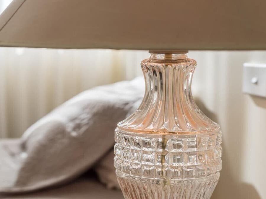 A clear glass table lamp with a beige lampshade on a bedside table.